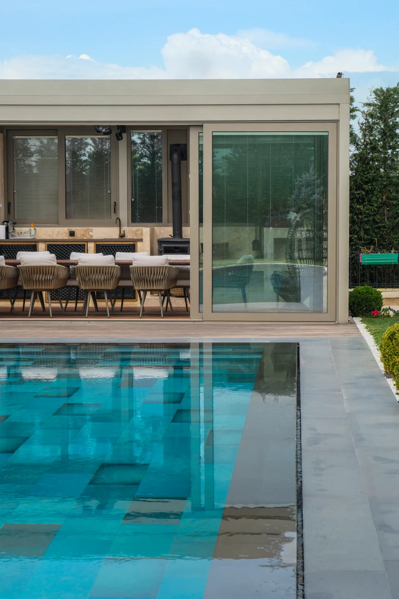 A residential glass-enclosed outdoor dining space beside a sunlit swimming pool.